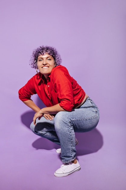 Modern woman with violet curly short hair in bright shirt, white sneakers and trousers bites her lip and squats on lilac.