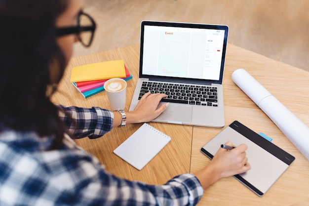 Free photo modern work image young brunette woman in black glasses from back working with laptop on table. creativity, graphic design, clever student, studying, freelancer.