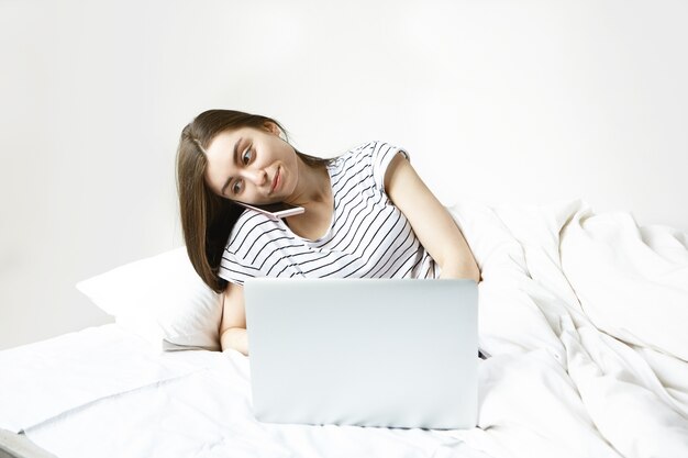 白いベッドリネンの上に横たわって、ポータブルコンピューターを使用して何かを入力し、電話で会話しながら縞模様のパジャマを着ている現代の若い20歳の女性