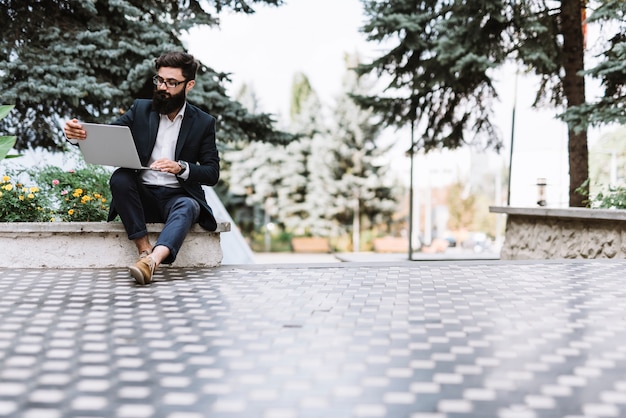 Modern young businessman sitting in the park looking at laptop