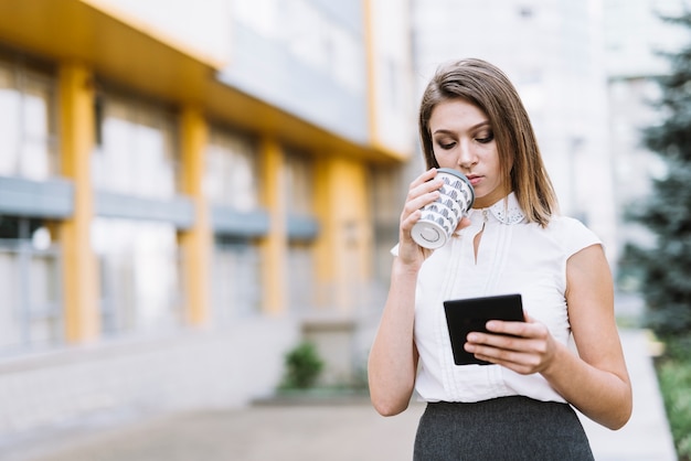 Modern young businesswoman drinking coffee while looking at smartphone