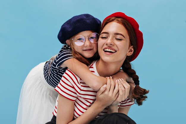 Modern young girl in eueglasses dark blue beret striped tshirt and white skirt hugging smiling charming elder sister with cool hairstyle