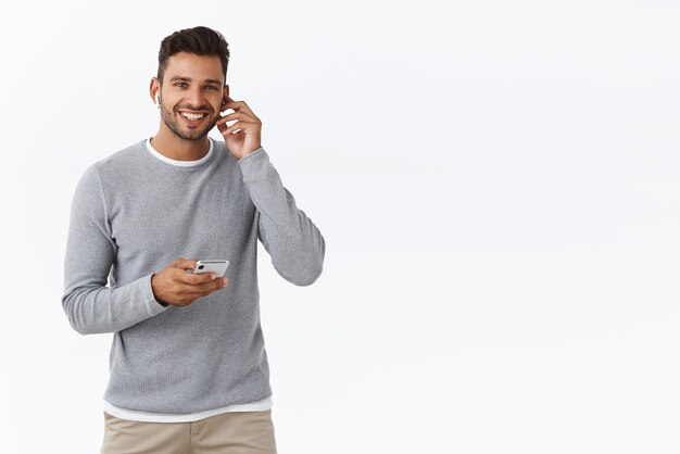 Modern youth and technology concept Happy smiling bearded male in grey sweater put wireless earphone in ear grinning delighted want call friend handsfree hold smartphone white background