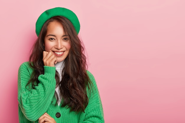 Modest adorable young woman smiles gently at camera, holds hand under chin, dressed in fashionable green outfit