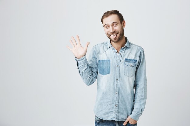 Modest cute bearded man say hi, waving hand in greeting, show tongue