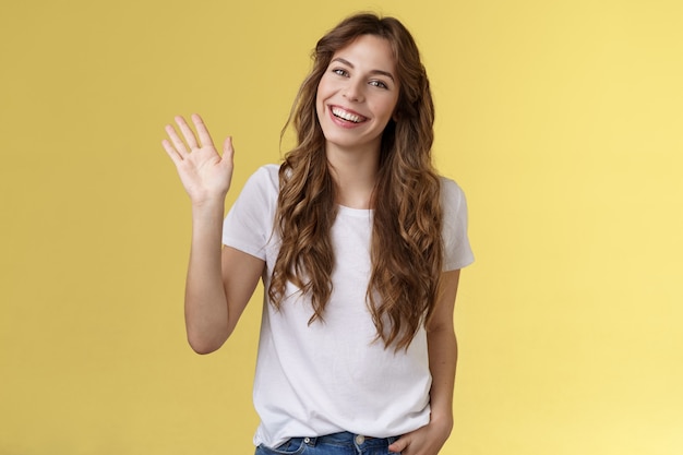 Modest cute friendly outgoing attractive european woman long curly haircut waving palm sociable conversation introduce herself smiling broadly say hi hello greeting coworker yellow background.