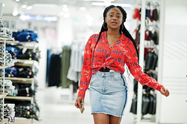 빨간 셔츠와 청바지 치마를 입은 모던한 아프리카계 미국인 여성이 옷가게에서 포즈를 취했습니다. 쇼핑할 시간입니다