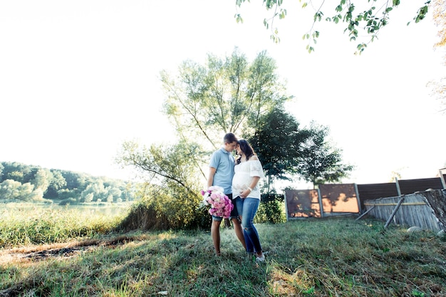 Modish expecting couple stands in warm embraces under green tree