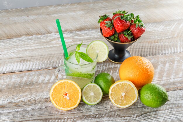 Mojito cocktail with limes, straw, oranges, lemon, strawberries, mint in a cup on wooden and grey surface, high angle view.