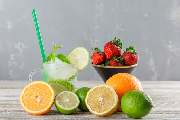 Mojito cocktail with limes, straw, oranges, lemon, strawberries, mint in a cup on wooden and plaster, side view.