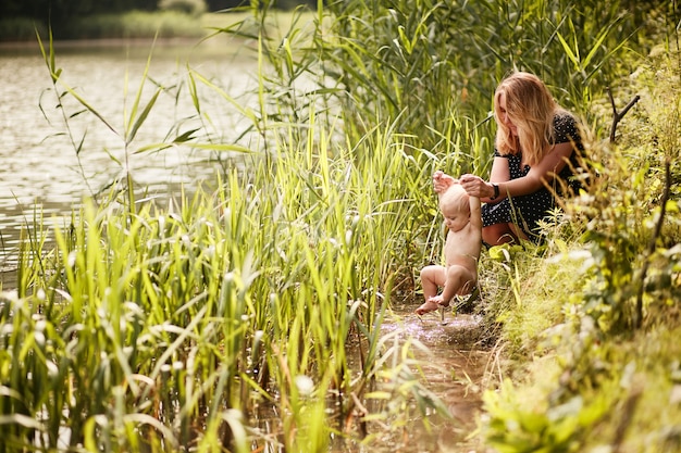 Mom bathes her little son in a river among tall green grass 