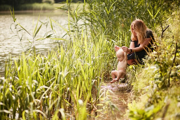 ママは背の高い緑の芝生の間で川で彼女の幼い息子を浴びる