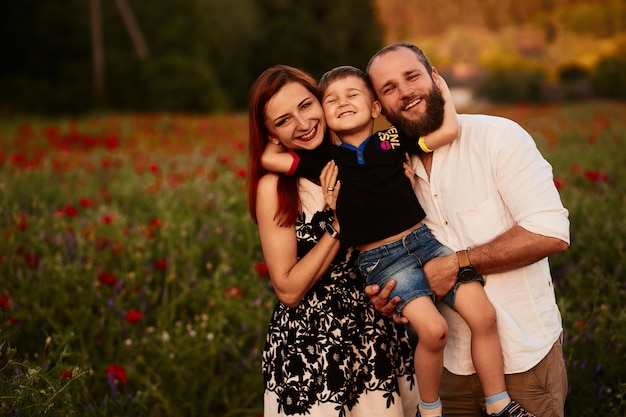 Mom and dad hold their little son on the arms standing on the green field with poppies