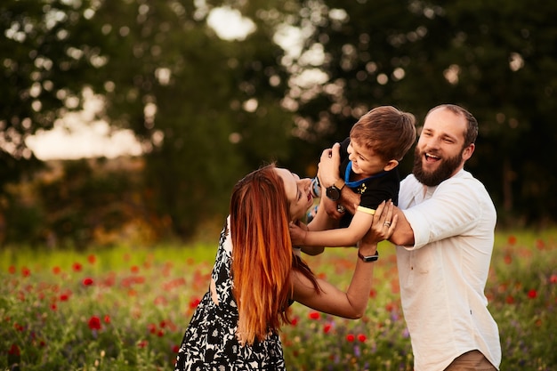 Foto gratuita mamma e papà reggono il loro piccolo figlio sulle braccia in piedi sul campo verde con i papaveri