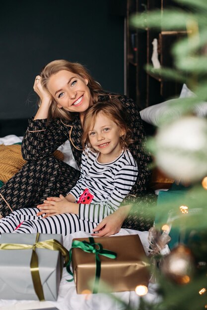 ママと娘は、クリスマス新年家族でクリスマス ツリーのそばのベッドでパジャマを着て座っています。