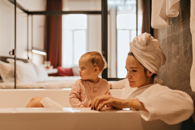 Mom and daughter are sitting in snow-white bath. Woman in towel on her head is holding baby.