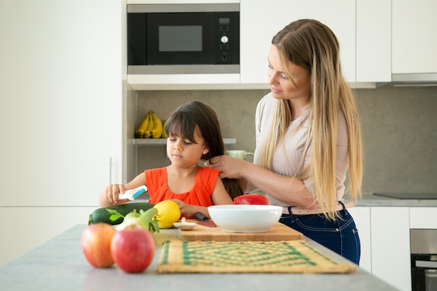 Mom and daughter together in kitchen. Mother braiding girls long hair, while kid peeling vegetables on counter. Family cooking concept