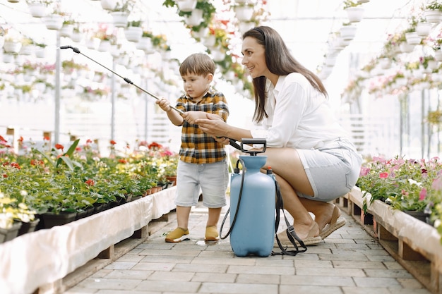 Mom and her son with a blue water sprayer in the greenhouse. Little toddler boy watering flowers in a greenhouse