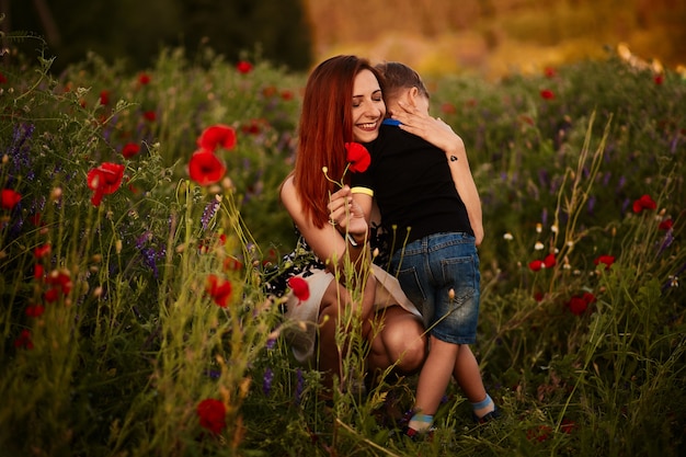 Mom holds charming little son standing on the green field with poppies