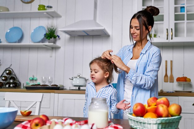 Mom is tying her daughter a ponytail and they will cook dinner in the kitchen at home.