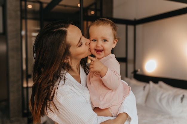 Mom kisses fair-haired child looking at camera with smile. Woman holds her daughter in her arms on background of white bed.