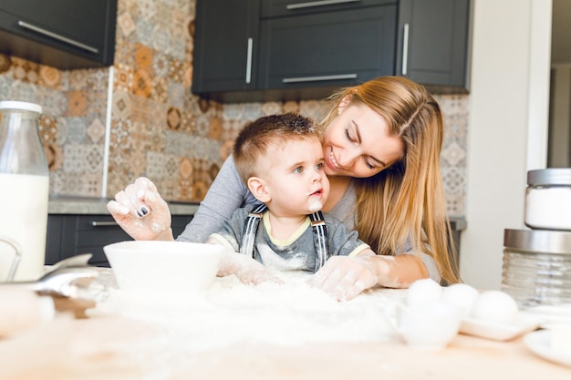 Mom playing with kid in the kitchen. Kitchen is done is dark colors and roustic style. Kid is covered in flour and looks funny. Table is made from light wood. Blender, milk, eggs, jars stand on table.