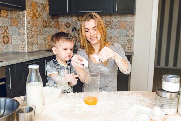 Mom playing with kid in the kitchen. Kitchen is done is dark colors and roustic style. Kid is covered in flour. Mom breaks egg into a jar. Blender, milk, eggs, jars lie on table.