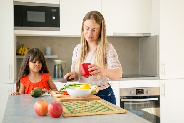 ママが娘に夕食にサラダを作る方法を示しています。少女と彼女の母親は台所のカウンターで野菜を切る。ミディアムショット、コピースペース。家族の料理のコンセプト