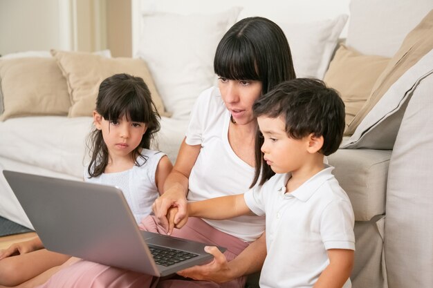 Mom teaching kids to use laptop, holding hand of little son and pressing keyboard button with boy finger.