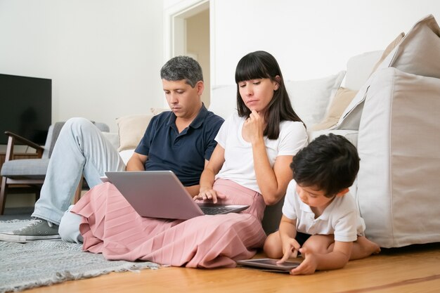 Mom with laptop watching little son using tablet, sitting on floor in living room by his parents.