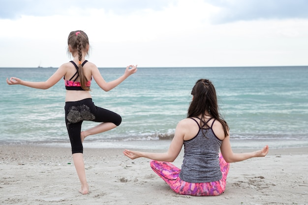 Мама с маленькой дочкой в спортивной одежде занимаются йогой на морском пляже, вид со спины. Семейные ценности и здоровый образ жизни.
