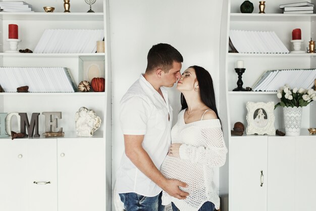 A moment before a kiss between expecting couple standing before white bookshelves 