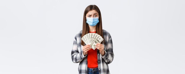 Money transfer investment covid19 pandemic and working from home concept Doubtful and confused young woman in medical mask holding cash and raising eyebrow suspicious or hesitant