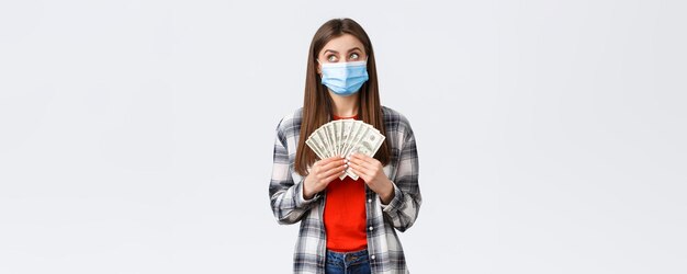 Money transfer investment covid19 pandemic and working from home concept Dreamy cute girl in medical mask holding cash dollars and look up imaging what buy dreaming of shopping or travelling