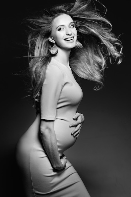 Monochrome portrait of beautiful happy pregnant woman in dress embracing belly and smiling Positivity and stylish female posing looking at camera Concept of pragnancy fashion Windy effect