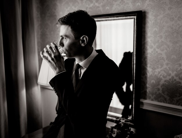 Monochrome portrait of handsome young man in room is drinking dressed in suit