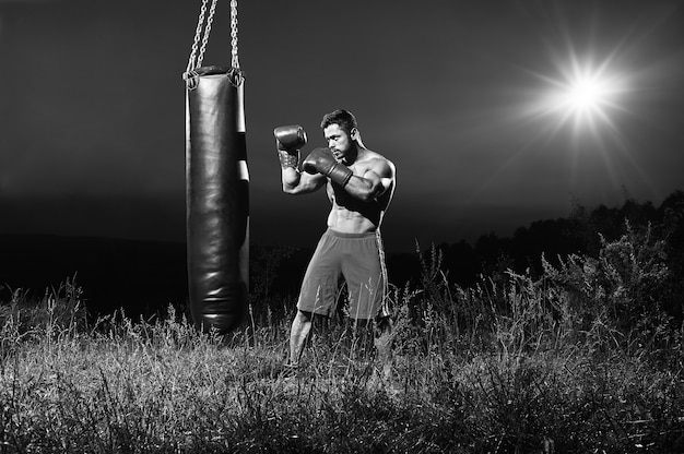 밤에 야외에서 훈련하는 잘생긴 젊은 근육질 남성 권투 선수의 흑백 초상화는 펀칭 백 카피스페이스 자연 단독 스포츠맨 경쟁적인 야심찬 강한 자신감을 가지고 연습합니다.