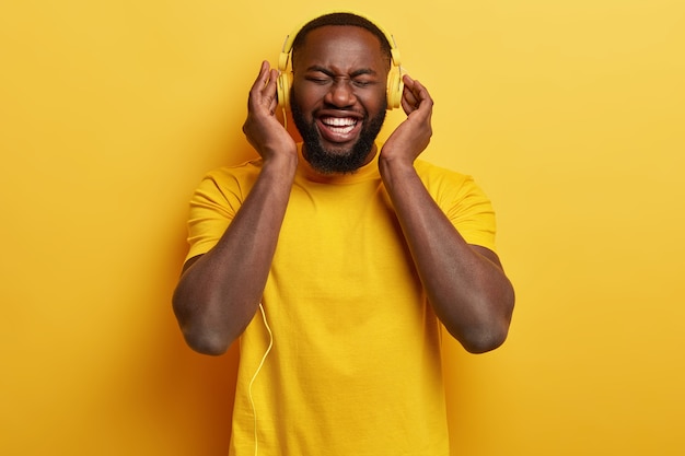 Monochrome shot of overjoyed pleased Afro American man enjoys perfect loud sound in new headphones, dressed in yellow t shirt, has free time, entertains him self with music. Happy expression.