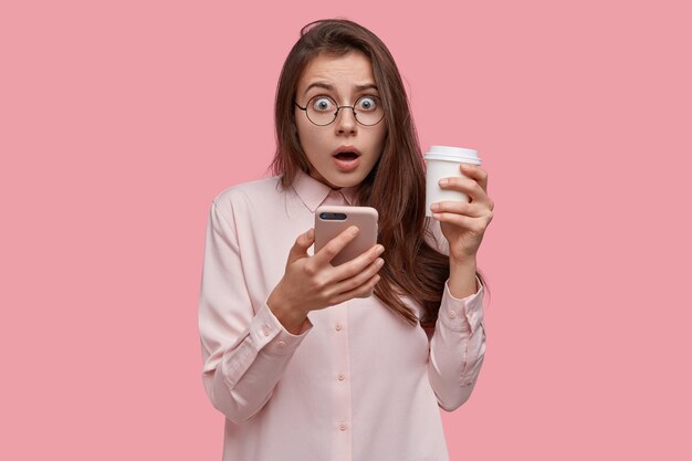 Monochrome shot of stupefied Caucasian woman opens mouth widely, reads incredible news on internet website, holds mobile phone, takeout coffee