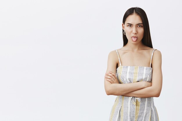 Moody and displeased attractive woman in cute dress, wrinkling nose and showing tongue, holding hands crossed on chest, expressing offence and dislike