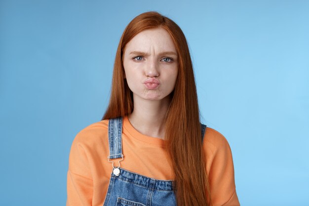 Moody displeased clingy girlfriend redhead blue eyes pouting sulking upset offended frowning making grimace showing attitude standing disappointed unsatisfied blue background, complaining