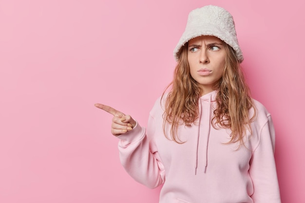 Moody dissatisfied young woman frowns face points idex finger away makes upset discontent grimace frowns from dislike makes negative opinion wears casual sweatshirt and panama isolated over pink wall