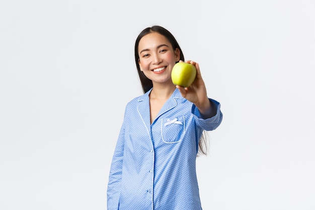 朝、アクティブで健康的なライフスタイルと家のコンセプト。青リンゴを見せて喜んでニヤリと笑うパジャマでフレンドリーな韓国の女の子を笑顔、健康を維持するために果物を食べることをお勧めします、白い背景。
