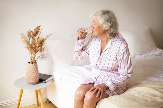 Morning rituals, lesiure, rest and bedtime concept. Attractive female pensioner with gray hair sitting on bed in cozy interior, holding mug, drinking water to make her digestive system work