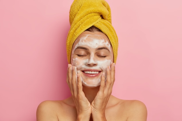 Morning routine and relaxing time concept. Pleased young woman washes face, cleans skin with soap, wears yellow towel on head, keeps eyes shut from pleasure