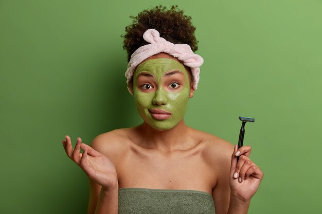 Morning skin care routine. Unaware woman shrugs shoulders with hesitation, applies moisturizing mask on face, holds razor for shaving, wrapped in bath towel, poses against vivid green wall