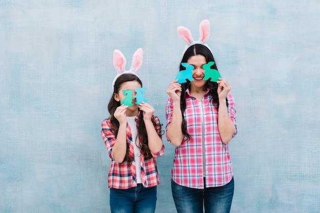 무료 사진 어머니와 딸이 파란색 벽에 눈 앞에서 종이 컷 아웃 토끼를 들고
