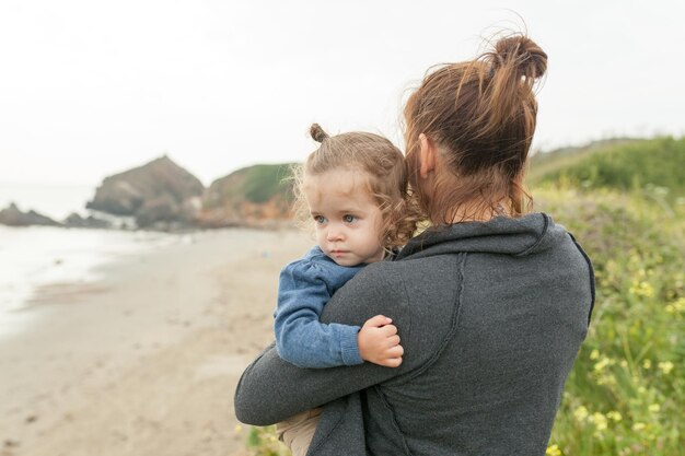 어머니와 작은 딸 서 있는 바위 바다 해변가 시즌. 한 아이가 함께 여행하는 백인 가족. 바다 풍경 배경 손에 어린 소녀 2 년을 들고 엄마