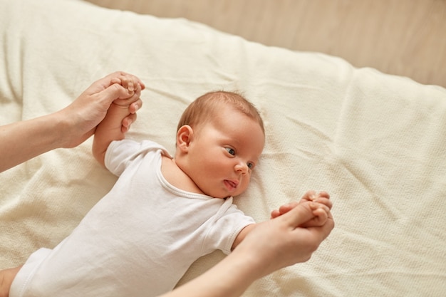 Mother and child in infant nursery doing exercises for baby's arms, kid looking away, wearing white bodysuit, lying on blanket, mommy holding kid's hands.