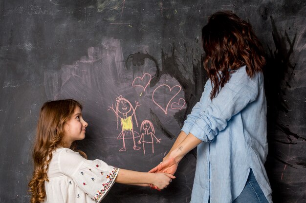 Mother and daughter holding hands near chalkboard with drawing 